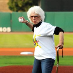 An elderly woman with a cane in hand prepares to through a baseball with a stern face.